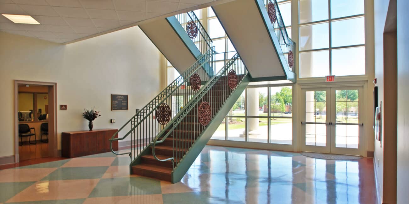 A bright, airy lobby with large windows, pastel tile floors, a decorative staircase, glass doors to the outside, and a reception desk.