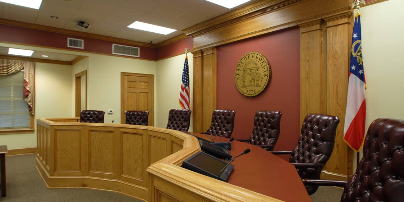 A courtroom with wood-paneled walls, leather chairs at a curved judge’s bench, desk microphones, and US and Georgian flags by a wall seal.
