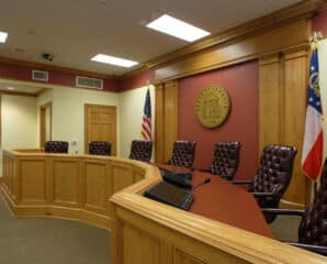 A courtroom with wood-paneled walls, leather chairs at a curved judge’s bench, desk microphones, and US and Georgian flags by a wall seal.
