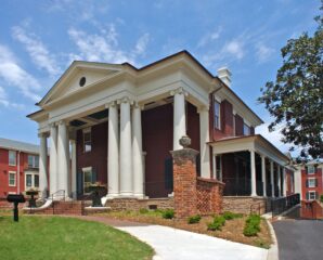 A large red-brick historic house with white columns and a triangular pediment, surrounded by lawns and nearby buildings under a blue sky.