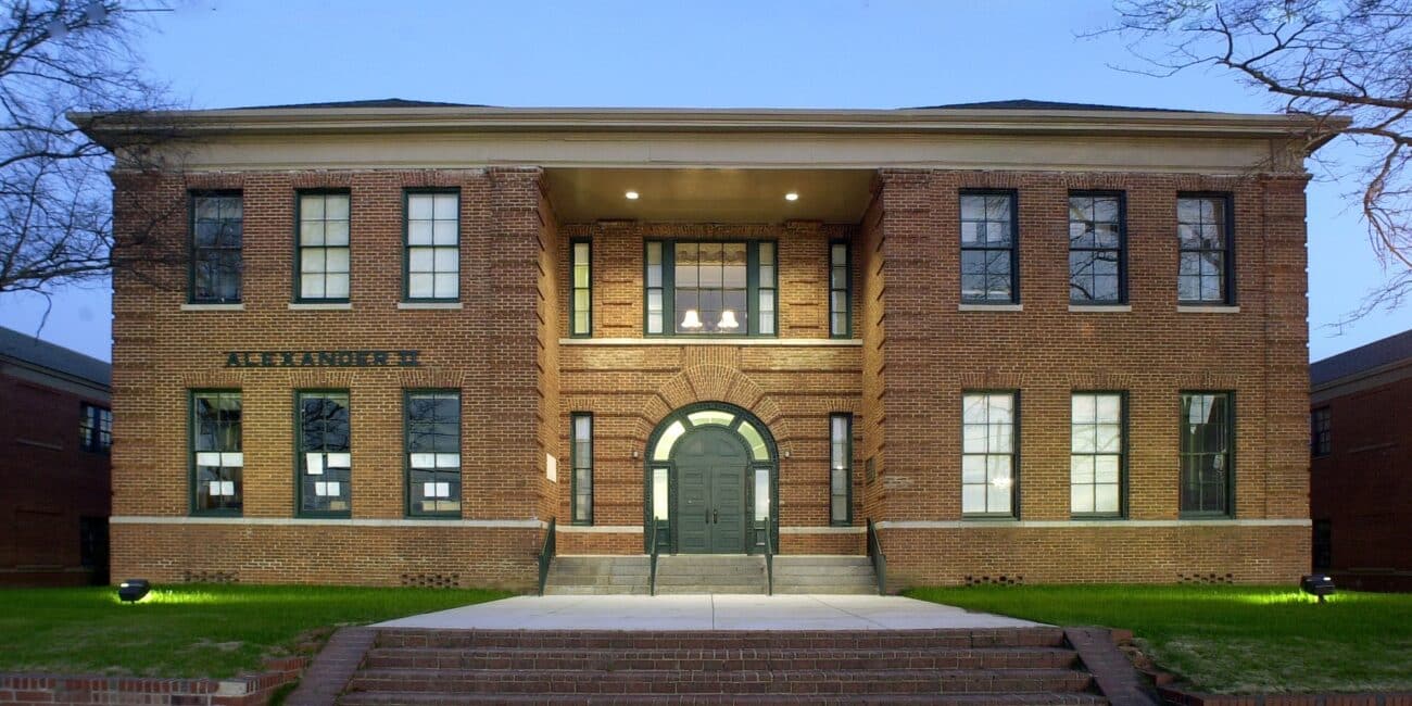 A two-story brick building with large windows, central arched doorway, ALEXANDER above the entrance, lit at dusk with trees and steps.