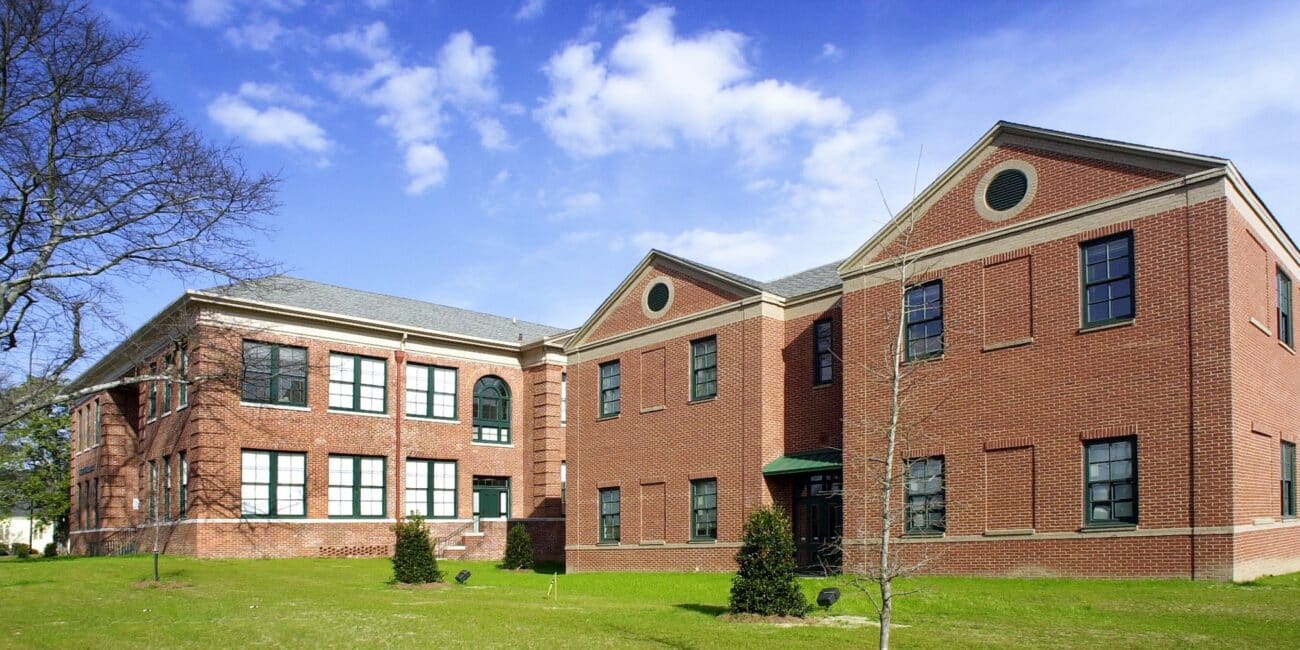 Large red-brick building with numerous windows, green grass and small trees around it, beneath a blue sky with scattered clouds.
