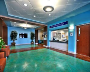 A brightly lit business center lobby with teal floors, blue walls, brown chairs, green plants, a reception desk, and a "Business Center" sign.