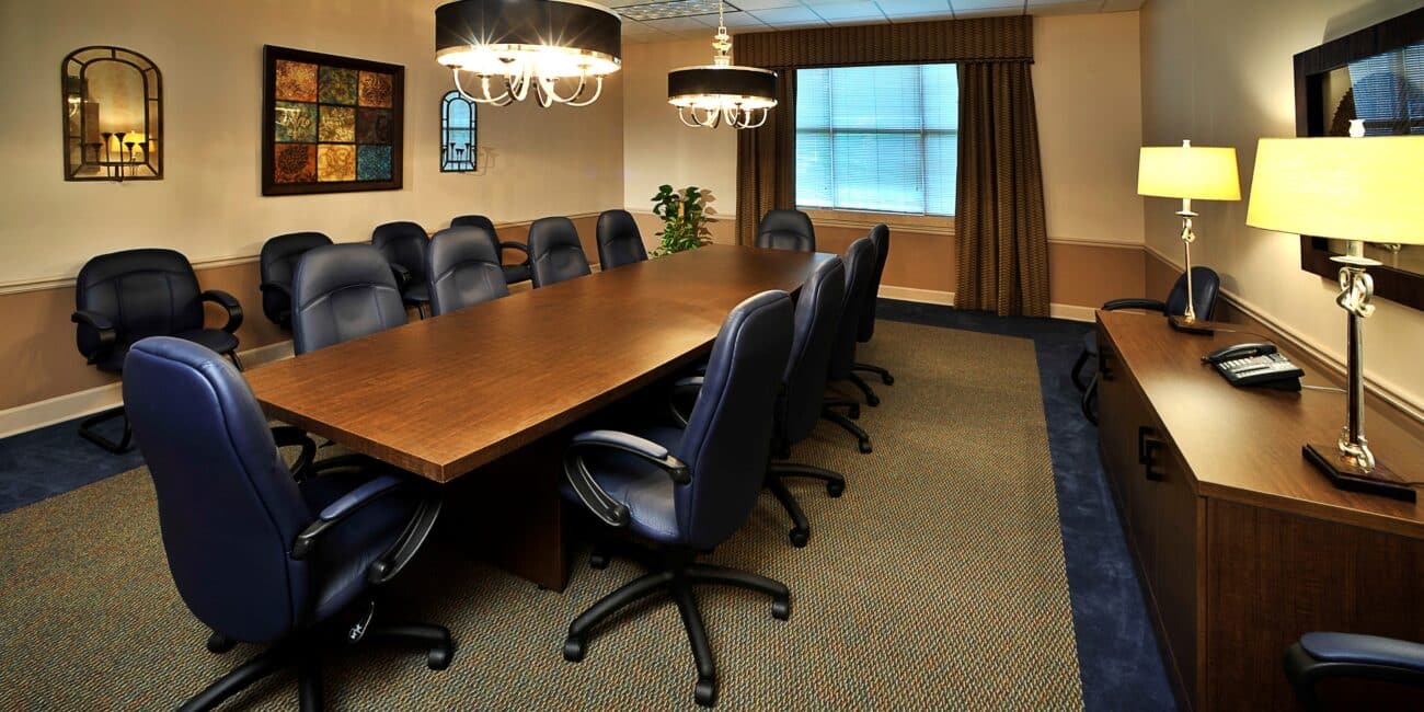 Modern conference room with a long wooden table, ten blue chairs, wall art, sideboard with lamps and phone, chandeliers, and closed blinds.