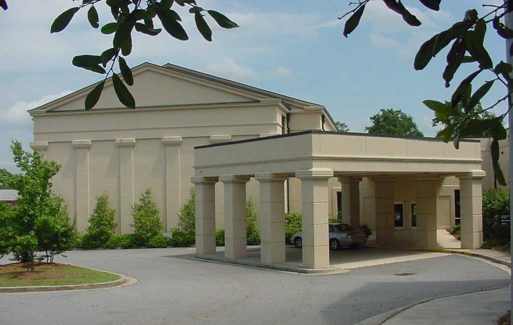 Beige building with tall columns and a covered driveway, greenery around, cloudy sky, seen through leafy branches in front.