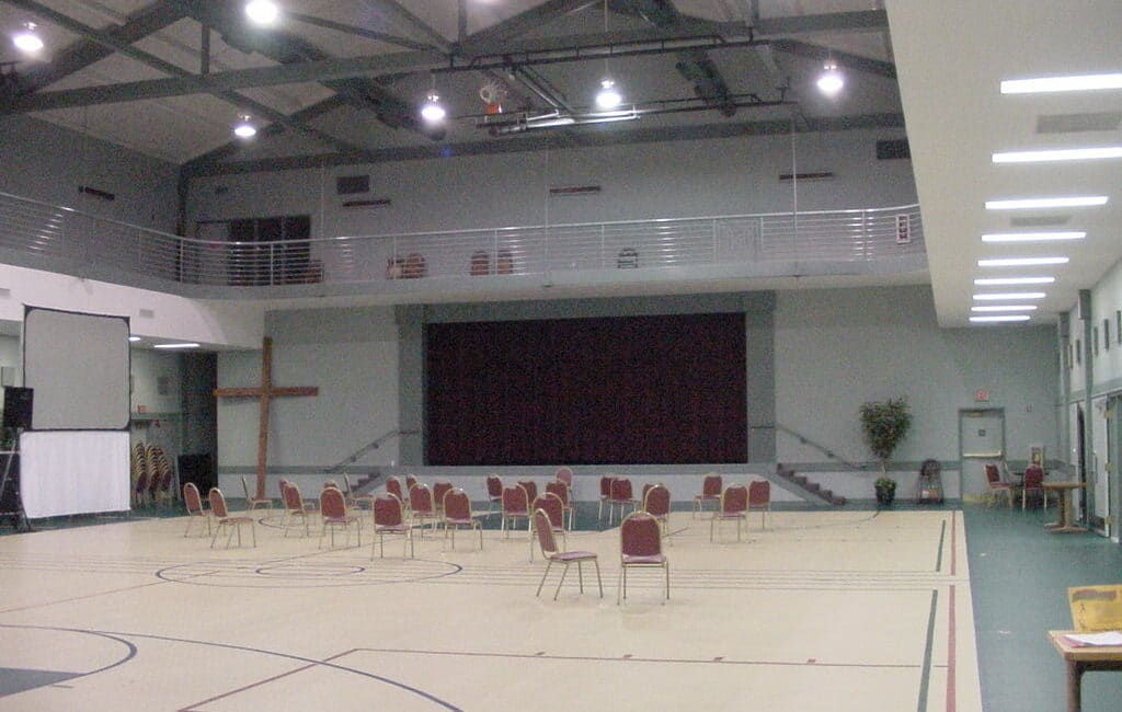 A spacious gym with red chairs, a stage and dark curtain, wooden cross, upper balcony, high ceilings, fluorescent lights, and court floor.
