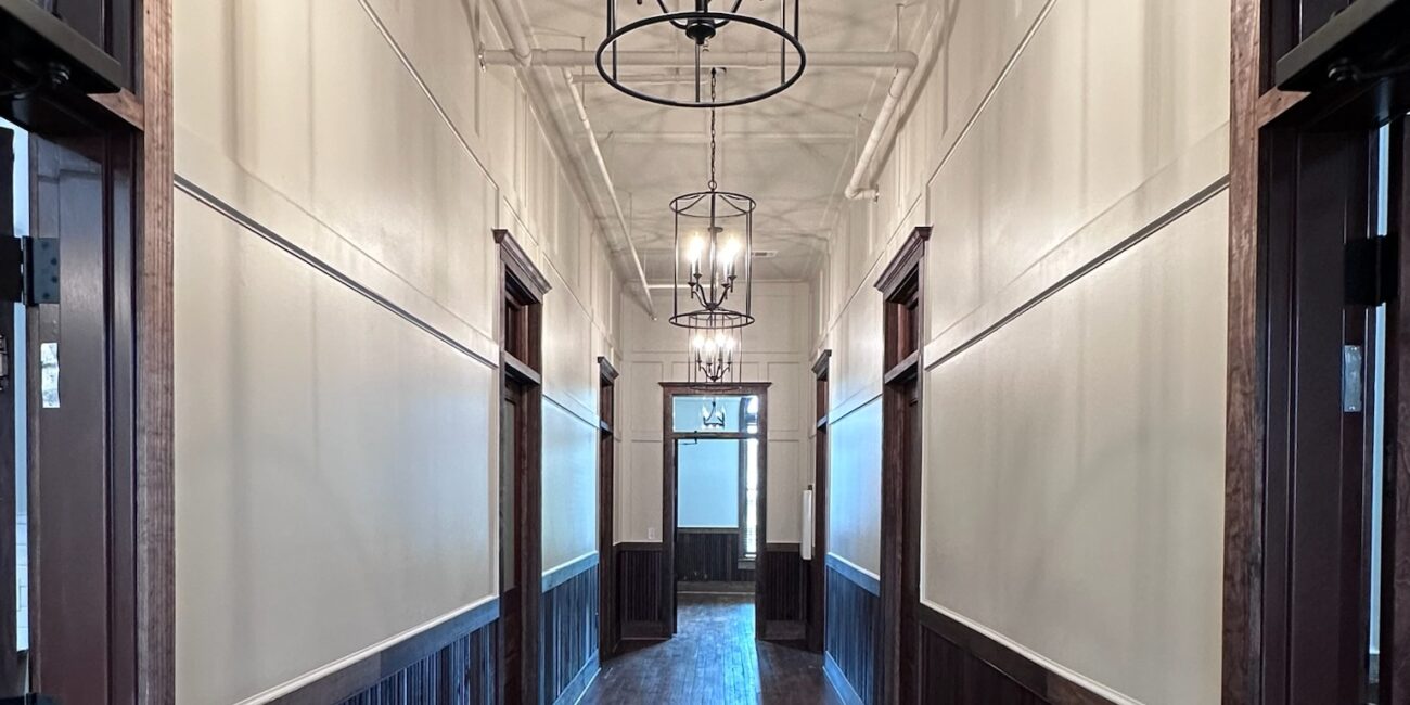 A long hallway with wood floors, dark paneling on lower walls, white upper walls, open doorways, and black chandeliers overhead.