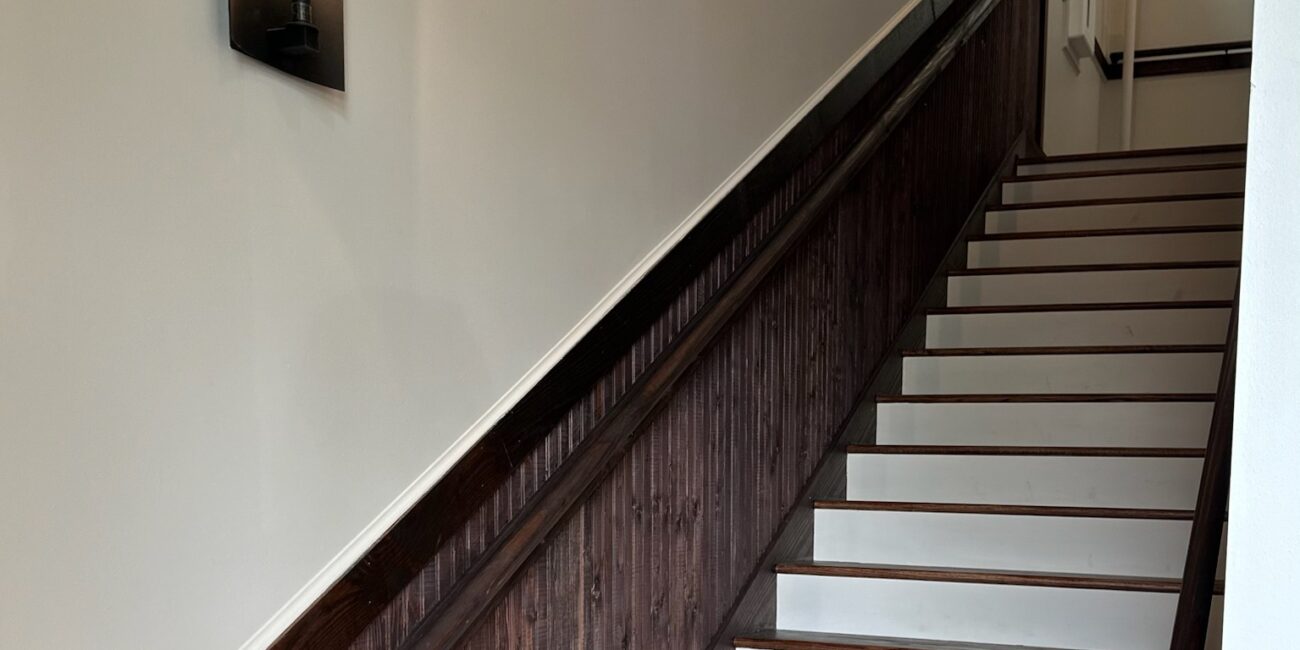 A staircase with dark wooden handrails and white steps ascends beside a beige wall lit by three sconces; window light at the top.