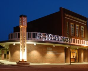 A brick building at night with a piano key motif over the entrance, lit sign Singing River Music, Visitors Center pillar, and address 450.