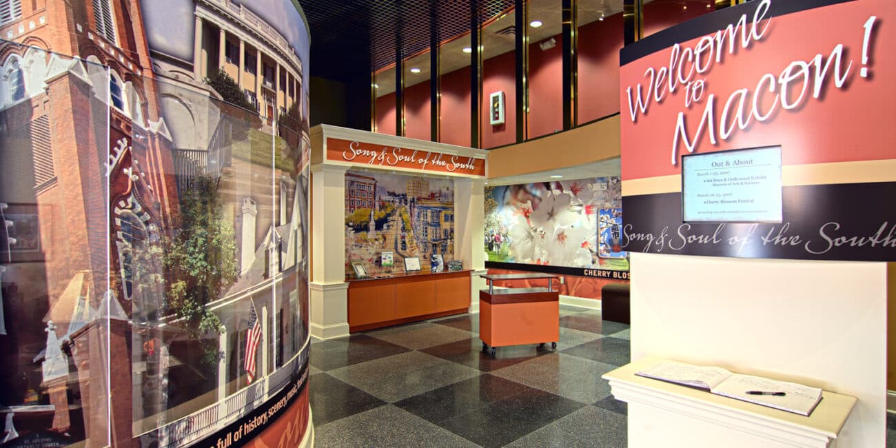 A brightly lit visitor center with displays and banners, a large sign reading Welcome to Macon, and images of local landmarks in Georgia.