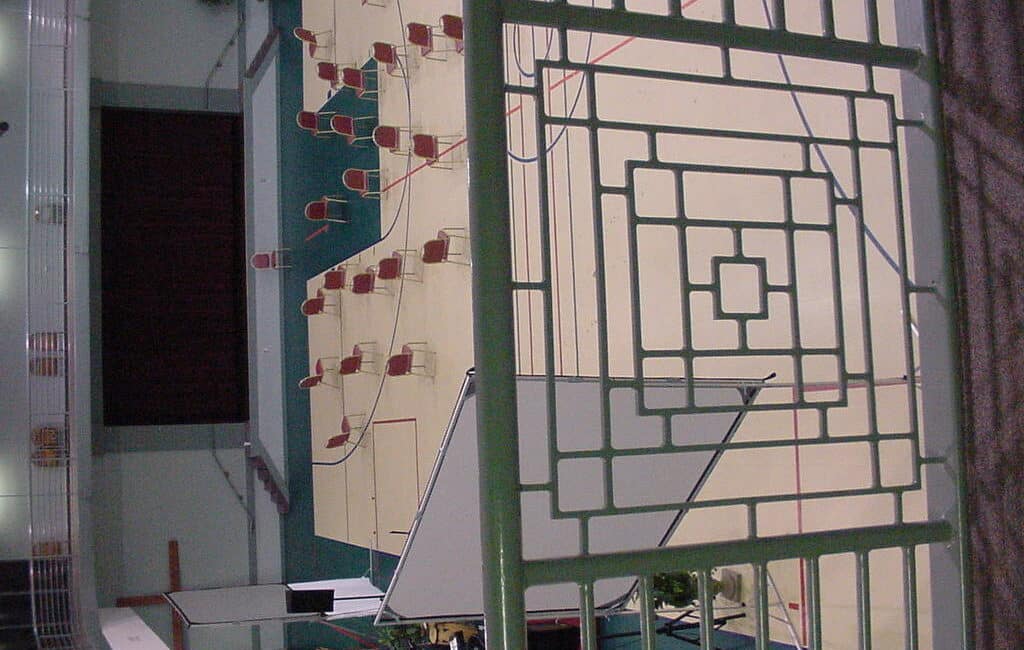 View from above of a gym or auditorium floor with scattered chairs facing a stage; foreground has a metal railing with geometric design.