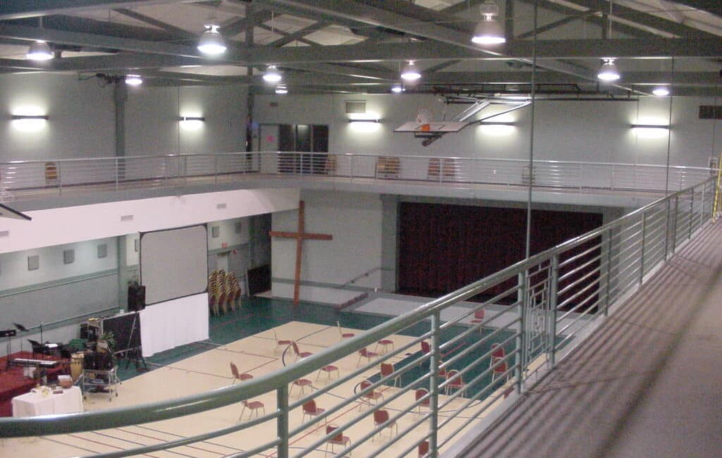 A gymnasium basketball court prepared for an event, with scattered chairs, large cross, screen, maroon-curtained stage, and upper walkway.
