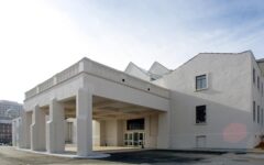 Modern white building with large covered entrance on tall pillars, simple facade, paved area outside, and clear blue sky above.