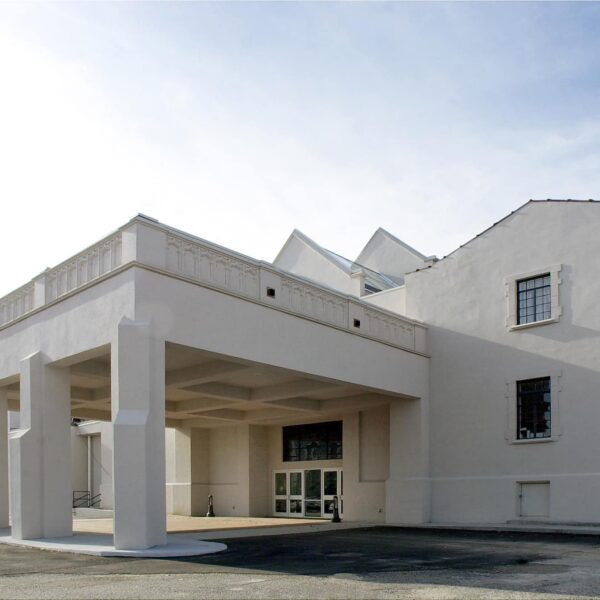 Modern white building with large covered entrance on tall pillars, simple facade, paved area outside, and clear blue sky above.