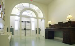 Bright hallway with double doors, arched windows, white bench left, ornate table with lit lamps right, framed pictures on wall.