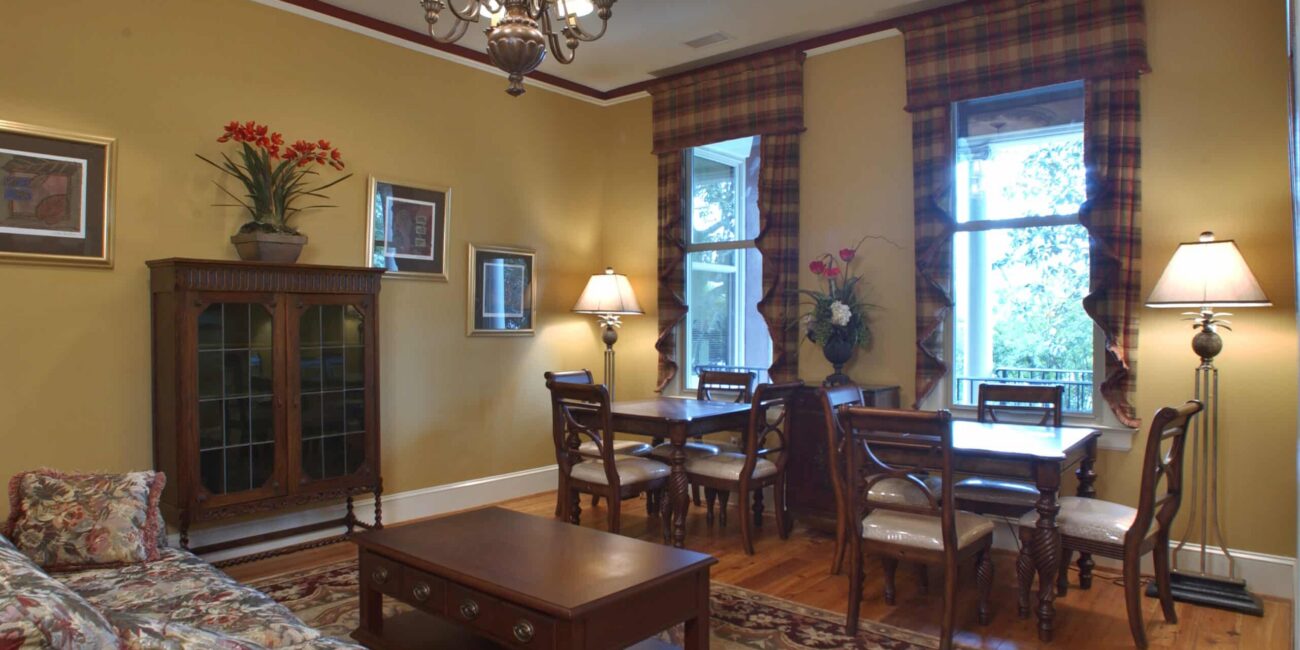 Traditional living and dining room with yellow walls, wood furniture, floral sofa, chandelier, large windows, and floral decor.