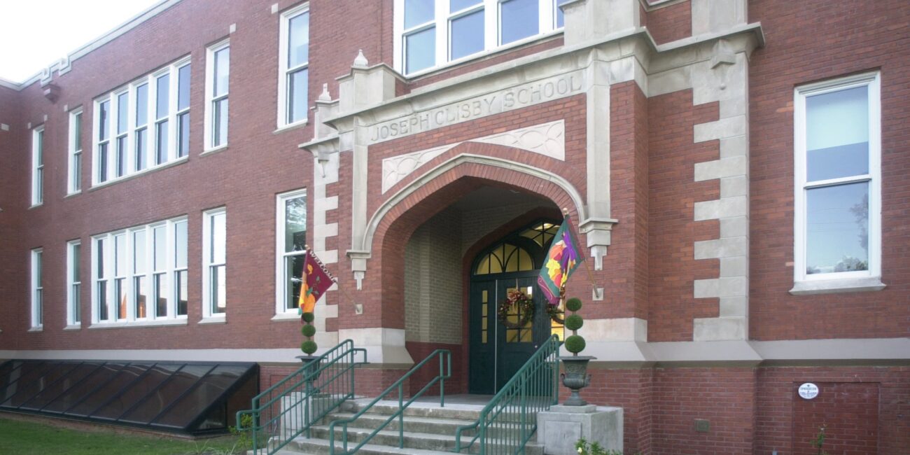 The entrance to a red-brick building labeled Joseph Clisby School, with arched stonework, green doors, flags, and green-railed stairs.