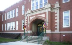 The entrance to a red-brick building labeled Joseph Clisby School, with arched stonework, green doors, flags, and green-railed stairs.