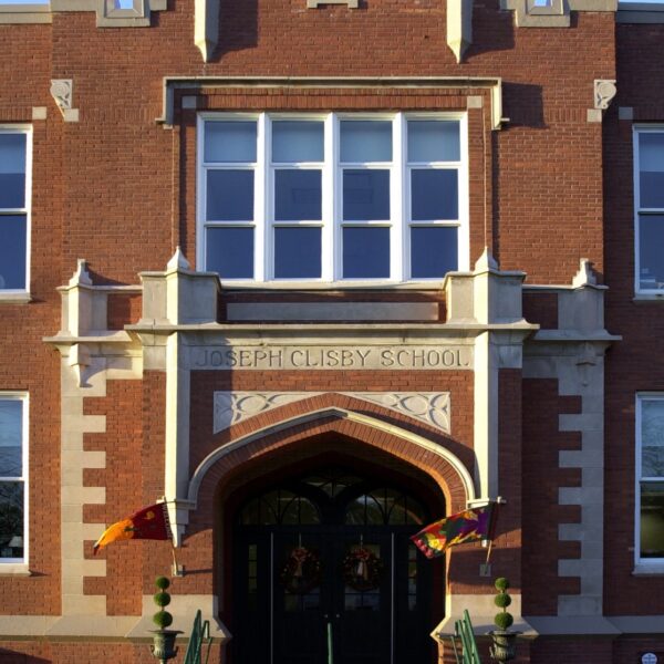 A red brick building with arched double doors, stone trim, and steps. Joseph Clisby School is engraved above, banners on flagpoles flank the entrance.