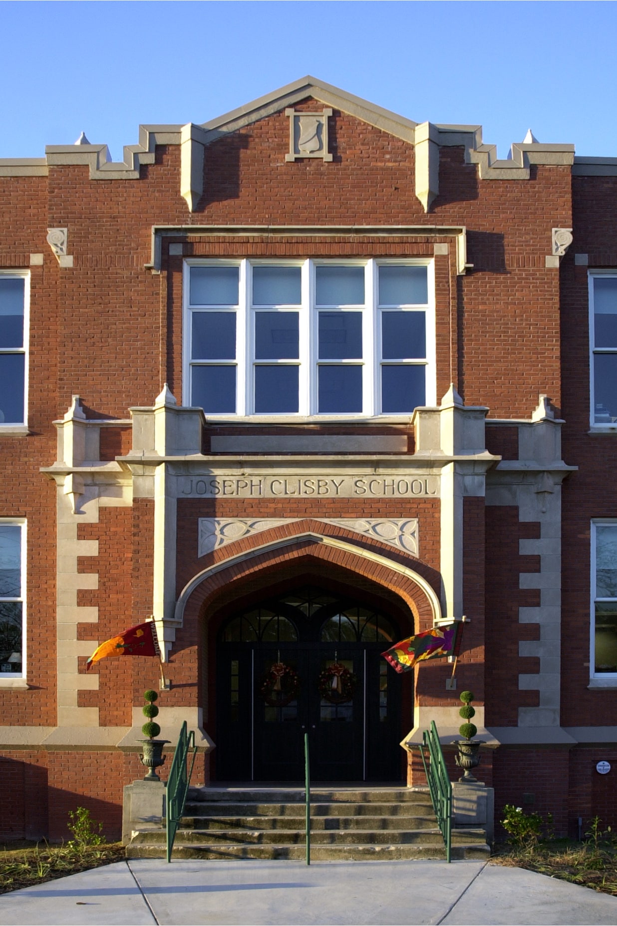 A red brick building with arched double doors, stone trim, and steps. Joseph Clisby School is engraved above, banners on flagpoles flank the entrance.