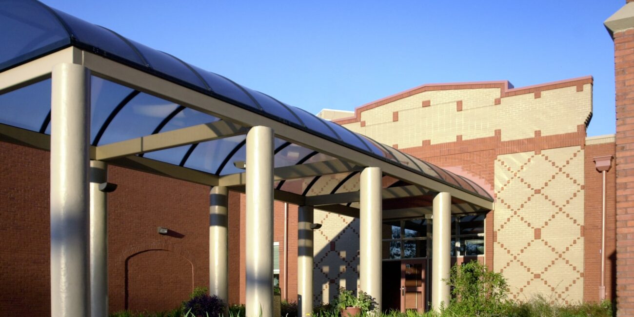 Modern building entrance with tan columns and glass canopy, light tan and red brick walls, geometric patterns, plants, blue sky.
