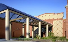 Modern building entrance with tan columns and glass canopy, light tan and red brick walls, geometric patterns, plants, blue sky.