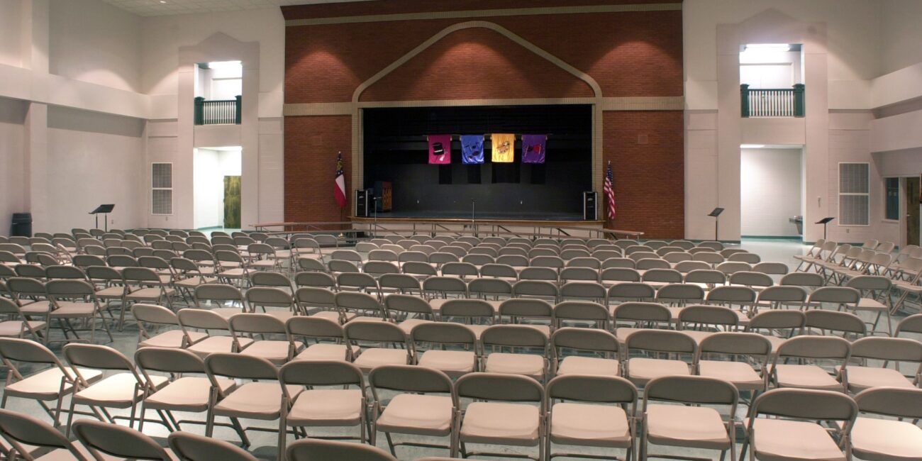 Rows of empty folding chairs face a stage in a bright auditorium with a brick backdrop, colorful banners, and flags on each side.