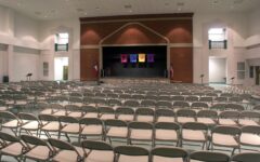 Rows of empty folding chairs face a stage in a bright auditorium with a brick backdrop, colorful banners, and flags on each side.