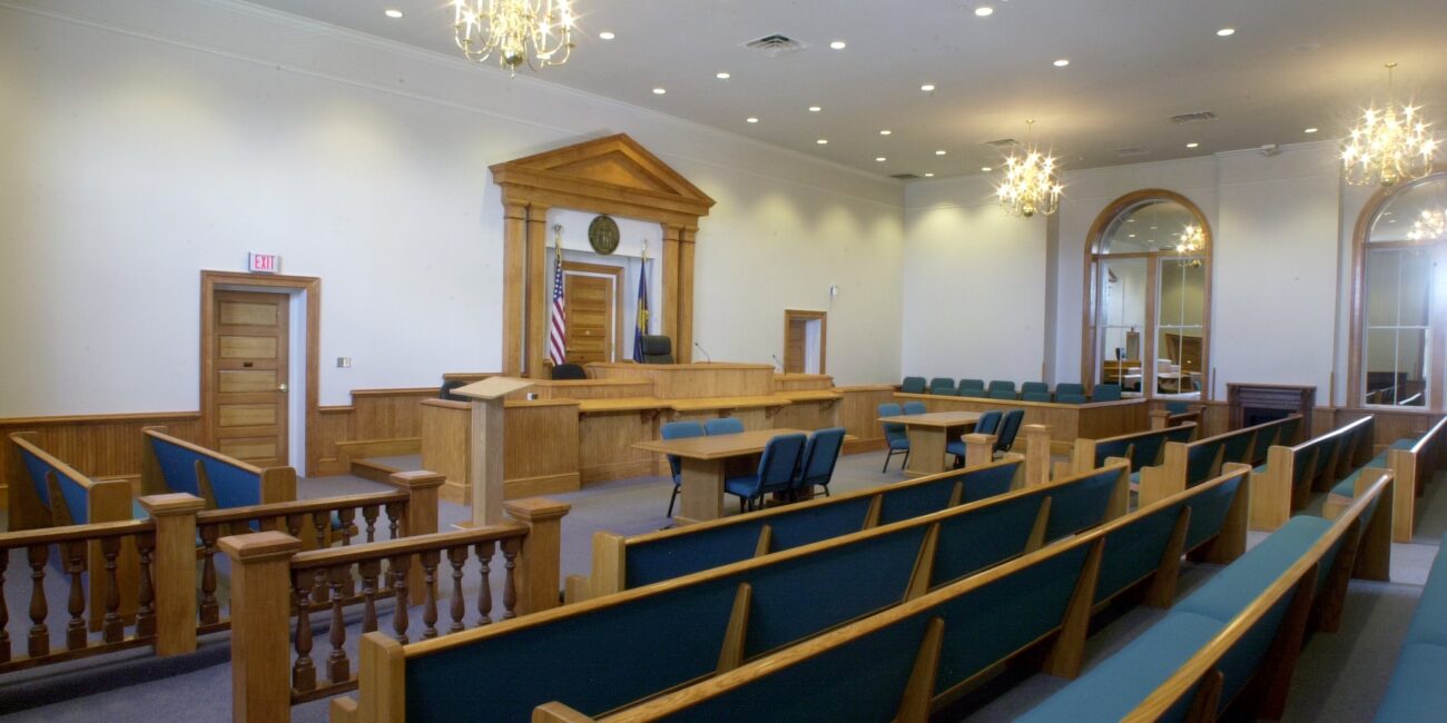 Spacious, empty courtroom with wooden benches, judge's bench, jury box, chandeliers overhead, and an American flag by the bench.