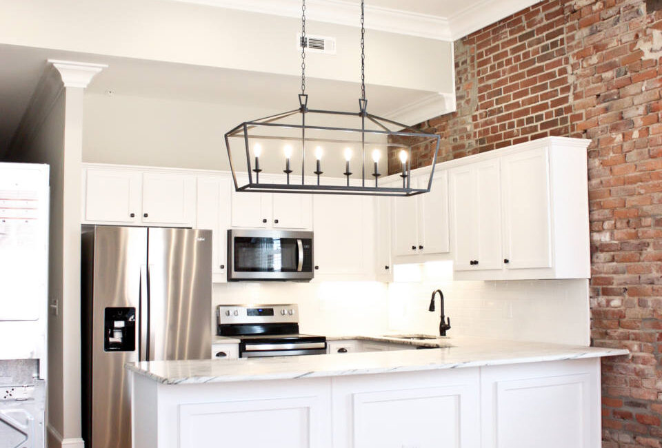 Modern kitchen featuring white cabinets, stainless appliances, brick accent wall, wood floors, and a bright minimalist design.