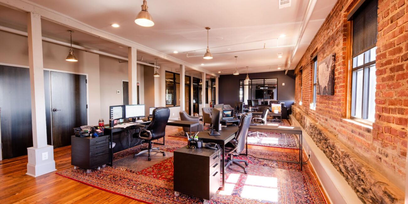 Modern open-plan office featuring brick walls, wood floors, big windows, exposed beams, and desks on colorful rugs in a cozy workspace.