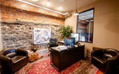 Cozy office with exposed brick and stone walls, desk with two monitors, black leather chairs, potted plant, colorful rug, artwork.