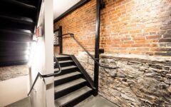 A staircase with black handrails and steps ascends beside an exposed brick and stone wall in a well-lit industrial-style interior.