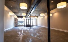 Spacious empty storefront with big windows, concrete floors, beige walls, exposed pipes, and pendant lights. Cars visible outside.