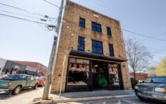 Three-story brick building with black-framed windows and a glass storefront on a corner, cars parked nearby under a clear sky.
