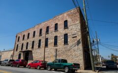 Historic three-story brick building with tall windows and parked cars including a green pickup, under blue sky and power lines.