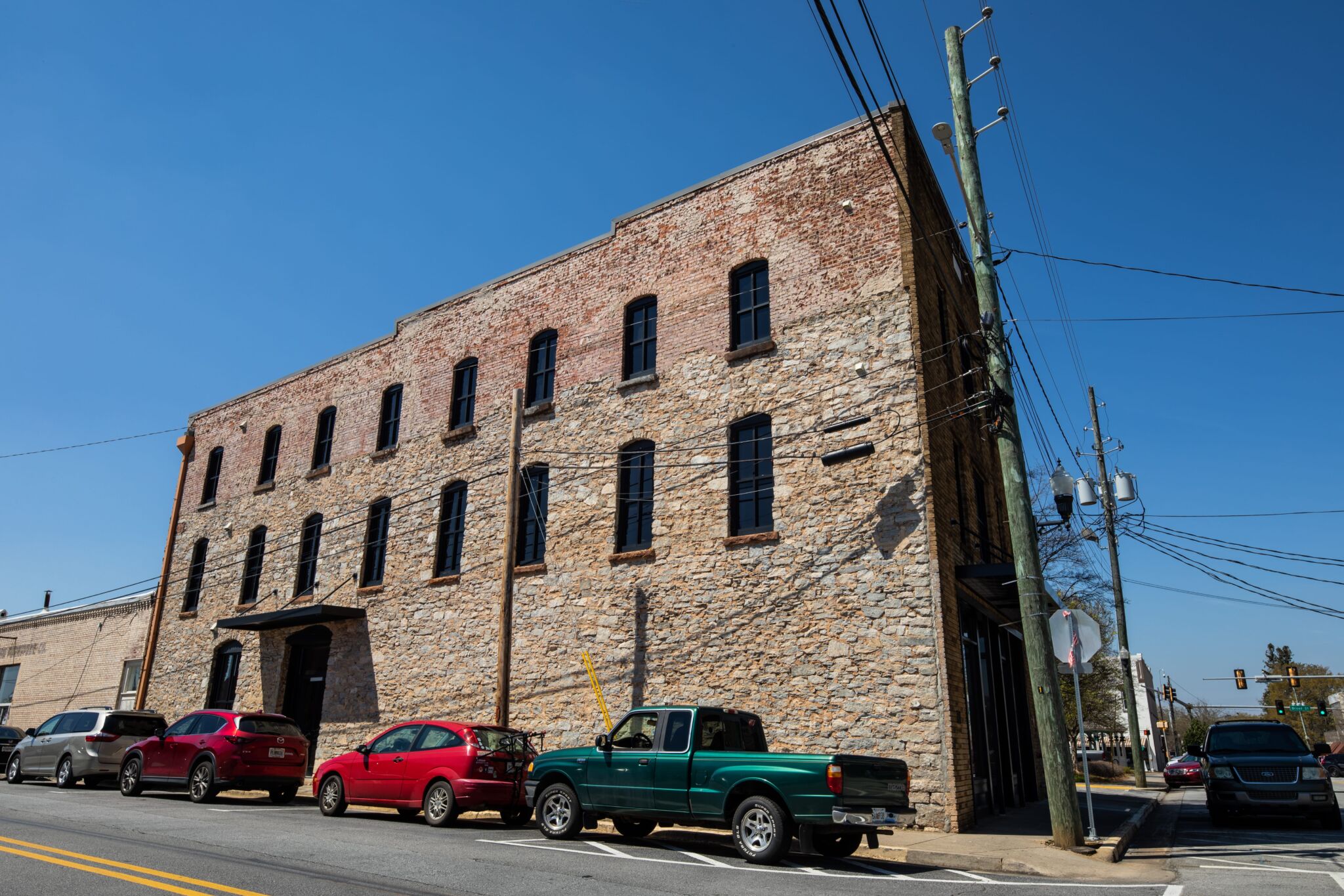 Historic three-story brick building with tall windows and parked cars including a green pickup, under blue sky and power lines.