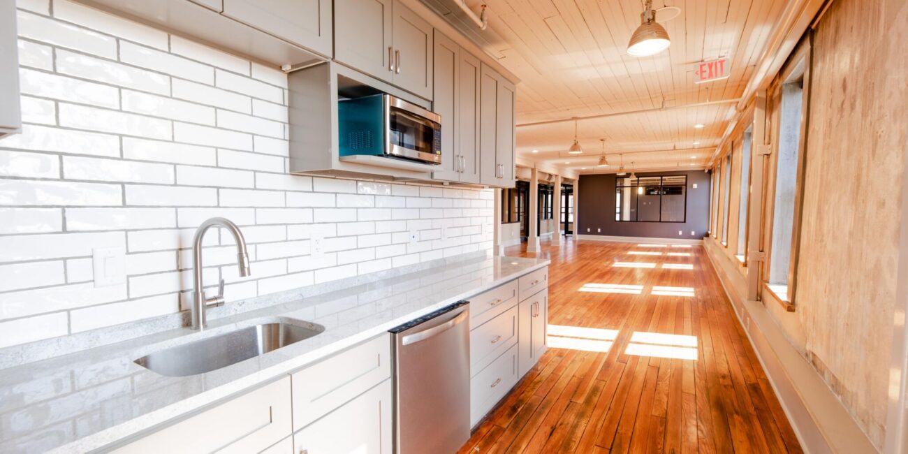 Modern kitchen featuring gray cabinets, stainless steel appliances, white subway tile backsplash, and sunlit wooden floor corridor.