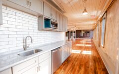 Modern kitchen featuring gray cabinets, stainless steel appliances, white subway tile backsplash, and sunlit wooden floor corridor.