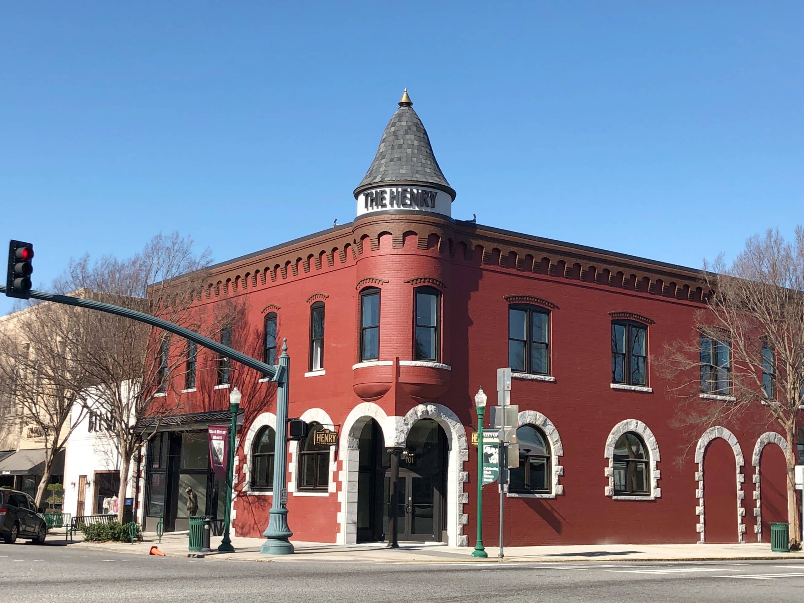 A two-story red brick building with arched windows and a cone-shaped turret displaying a sign that reads THE HENRY. The building sits on a street corner with a traffic light and street signs nearby.