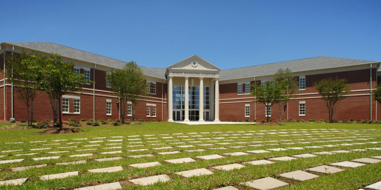 A red-brick academic building features tall white columns, large windows, and a stone walkway across a green lawn under blue sky.