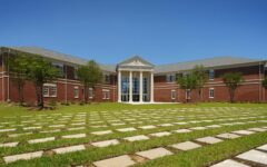 A red-brick academic building features tall white columns, large windows, and a stone walkway across a green lawn under blue sky.