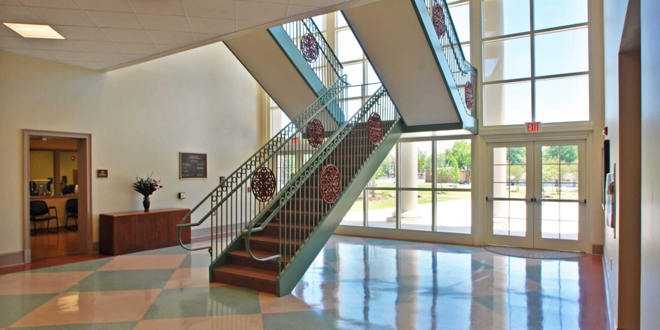 Bright, spacious lobby with large windows, colored tile floor, central staircase, double glass doors, and a reception desk in back.