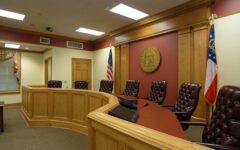 A courtroom with five leather chairs behind a wooden bench, microphones, two flags, and the Georgia seal. Wood paneling, carpeted floor.