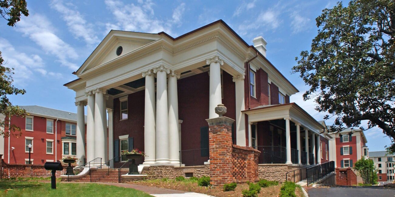 A large historic red-brick building with white columns and a triangular pediment stands under a blue sky, lawn and walkway around.