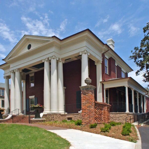 A large historic red-brick building with white columns and a triangular pediment stands under a blue sky, lawn and walkway around.