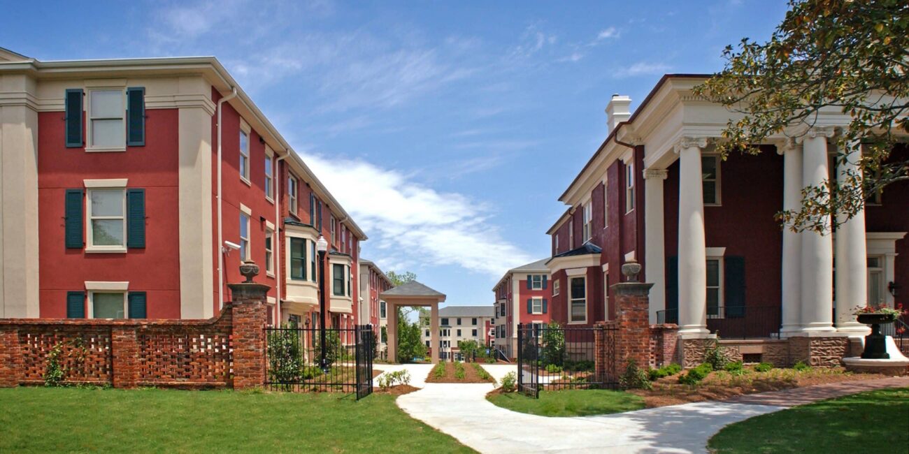 A curved path runs between red-brick colonial buildings with white columns, leading to more buildings under a blue sky with clouds.