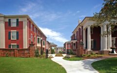 A curved path runs between red-brick colonial buildings with white columns, leading to more buildings under a blue sky with clouds.