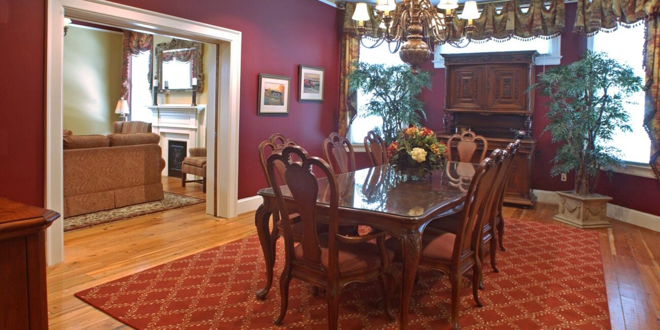 Traditional dining room with a wooden table, eight chairs, red rug and walls, chandelier, curtains, plants, and living room view.