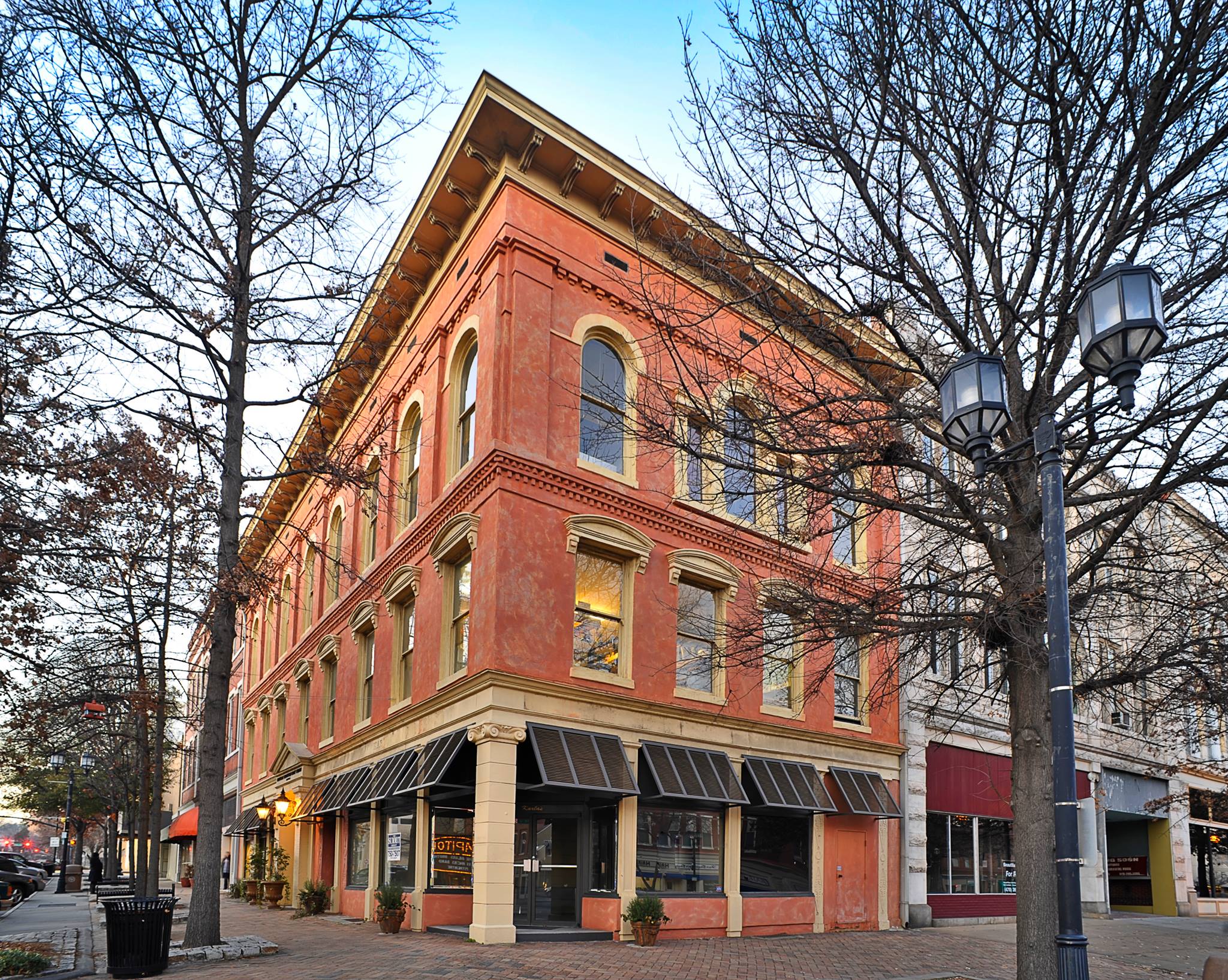 Three-story red-brick building with arched windows and black awnings on a tree-lined corner, bare trees and lamppost in front.